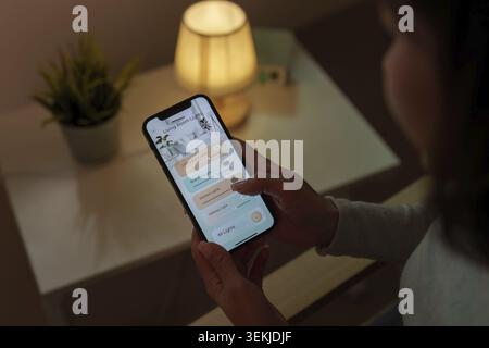 A person using a smartphone to control a smart home lighting system, shown in a cozy room with a lamp and plant. Concept of IoT and home automation Stock Photo