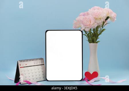 A Mother's Day-themed setup featuring a calendar, pink carnations in a vase, a blank tablet screen, and a red heart on a blue background Stock Photo