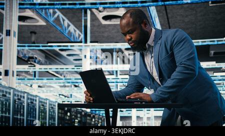 Programmer at server farm workplace desk using laptop, doing network management. Data center IT expert overseeing IT infrastructure supercomputers with software, typing on notebook keyboard, camera A Stock Photo