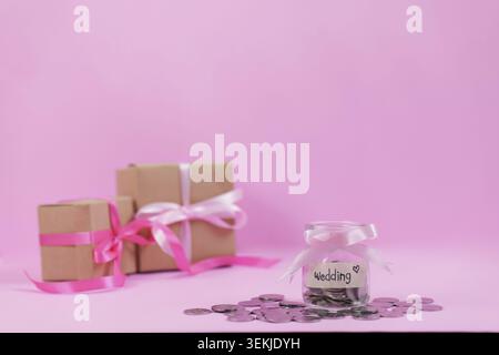 A jar labeled 'Wedding' filled with coins, surrounded by gift boxes on a pink background, symbolizing saving for wedding expenses Stock Photo