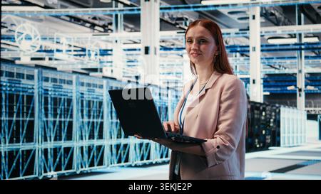 Happy woman in server room using notebook to enhance operational effectiveness of infrastructure. Joyous female engineer in data center using laptop to monitor rigs metrics, camera A Stock Photo