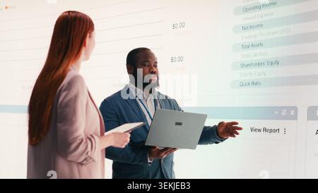 Businesspeople use laptop to analyze economic data graphs on dashboard, brainstorming. Teamworking colleagues look at charts projected on digital wall, verifying company financial statements, camera B Stock Photo