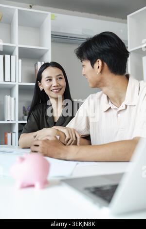 A young couple smiling and discussing financial planning and savings at home, with a pink piggy bank on the table Stock Photo