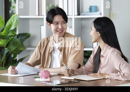 A young couple sits at a desk in their home, planning their finances with documents and a piggy bank, smiling and discussing their budget Stock Photo