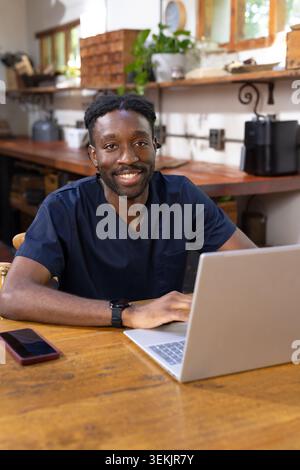 African American man sitting at wooden table wearing navy scrubs using laptop and smartphone. Workspace, healthcare, professional, cozy, sunlit, plant Stock Photo