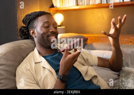 African man reclining on gray couch at home holding smartphone wearing beige shirt and smartwatch. Sofa, warm, cozy, ambient, lifestyle, casual, relax Stock Photo