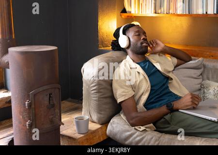 African American man reclining on light-gray leather sofa in cozy living room, wearing headphones. Snug, warm, rustic, ambient, home, lounge, bookshel Stock Photo