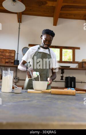 African man wearing green apron pouring milk into mixing bowl at kitchen counter with whisk Stock Photo