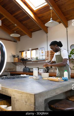 African American man in green apron cracking egg into mixing bowl on concrete island, copy space Stock Photo