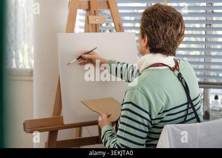 Rear view of a mature woman in a striped sweater holding a wooden palette while painting on a large white canvas on an easel in a bright home studio n Stock Photo