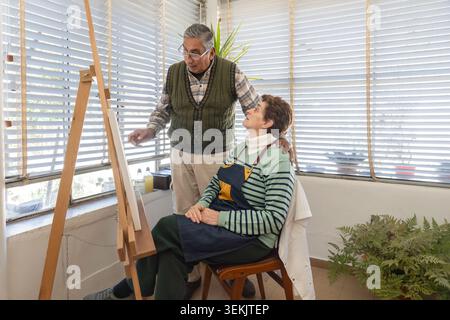 Senior couple sharing a cheerful conversation beside an easel in a bright home art studio. Stock Photo