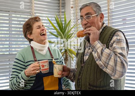 A senior woman bursts out laughing while holding two colorful ceramic mugs as her husband eats a croissant beside her, both sharing a joyful break in Stock Photo