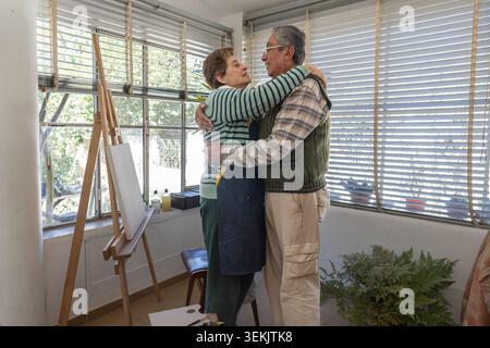 Happy older couple embraces warmly beside a wooden easel in a sunlit room, sharing an affectionate and playful moment during a painting session. Stock Photo