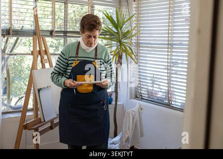 Senior female artist standing in a bright home studio holding a watercolor palette and brush beside an easel, expressing creativity, focus and active Stock Photo