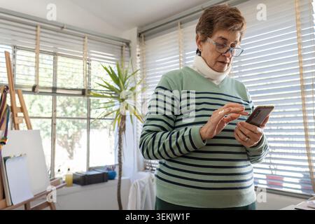 Older woman wearing blue reading glasses checks her smartphone in a bright home studio, with a wooden easel and art supplies visible behind her. Stock Photo