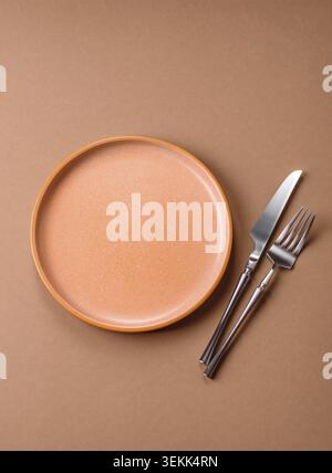 An empty round ceramic plate, knife, and fork as a dining table setting. An empty plate as a backdrop for your design Stock Photo