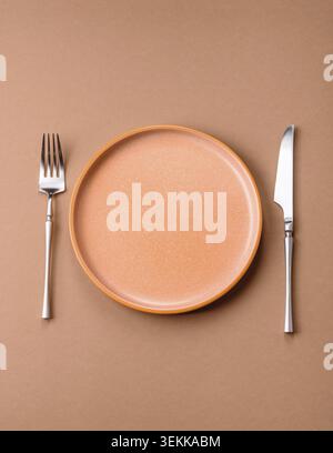 An empty round ceramic plate, knife, and fork as a dining table setting. An empty plate as a backdrop for your design Stock Photo