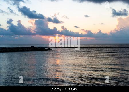 Sunrise over the ocean with dramatic storm clouds illuminated by golden sunlight, crepuscular rays spreading across the sky above calm tropical waters Stock Photo