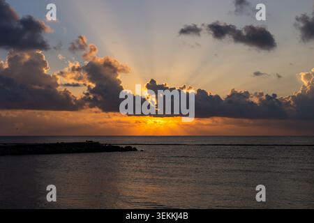 Sunrise over the ocean with dramatic storm clouds illuminated by golden sunlight, crepuscular rays spreading across the sky above calm tropical waters Stock Photo