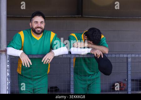 Two male baseball teammates wearing green jerseys leaning on railing in dugout, holding black cap Stock Photo