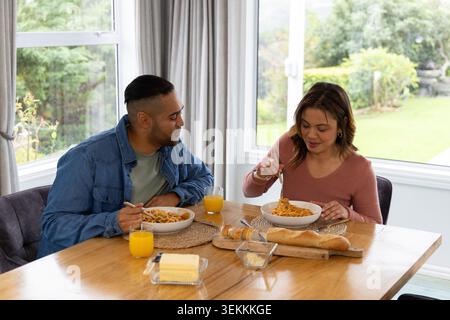Mid-adult couple sitting at dining table by window twirling spaghetti with baguette and juice Stock Photo