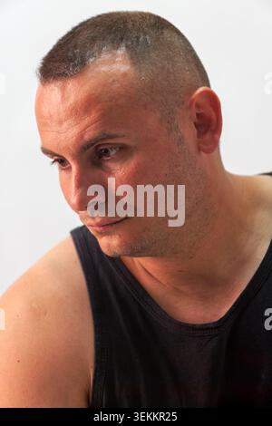 Close-up studio portrait of a man with a short buzz cut hairstyle wearing a black tank top and looking downward against a neutral grey studio wall. Stock Photo