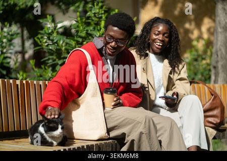 Happy african american friends man woman enjoying company of stray cat while resting on park bench Stock Photo