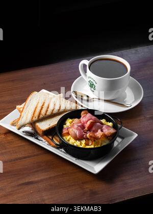 Cozy breakfast spread on dark wood table. Black coffee steams beside toasted bread. Fried eggs with bacon nestled in small bowl. Golden spoon rests on Stock Photo