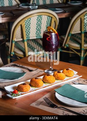 Warm wooden table holds appetizing croquettes. Glass of dark sangria glows beside them. Chairs with woven patterns frame the setting. Tableware rests Stock Photo