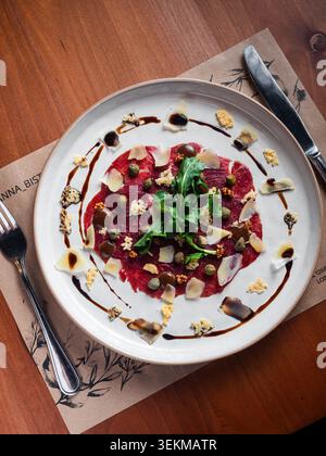 Delicate dish rests on rustic table setting. Fresh greens crown vibrant red center. Capers, nuts, and drizzles add texture and flavor. Simple elegance Stock Photo