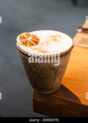 Warm drink sits in rustic ceramic cup. Citrus slice adorns creamy foam top. Cup rests on wooden table edge. Soft lighting highlights rich textures. Co Stock Photo