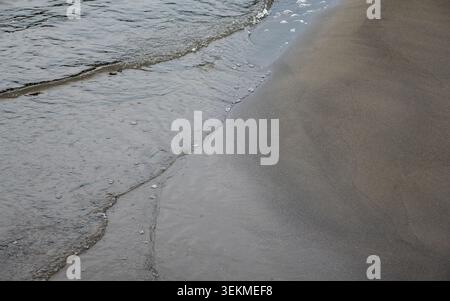 High angle view of calm water ripples and bubbles on dark volcanic sand at the waters edge Stock Photo