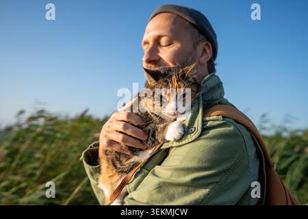 Domestic calico cat wearing leash, walking outside for first time, sits on man shoulder. Fluffy tabby feline carefully breathes fresh air without gett Stock Photo