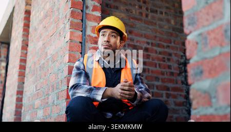 Man, architect or thinking with helmet in building site for construction break or rest on brick wall. Male person, civil engineer or thoughtful Stock Photo