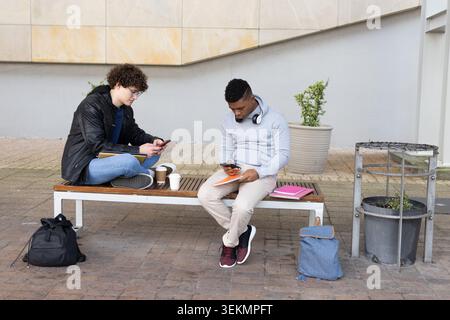 Diverse college males sitting on bench at campus courtyard using smartphones and notebooks Stock Photo
