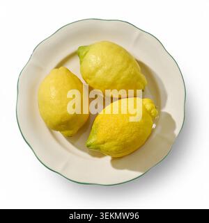 Overhead flat lay studio shot of three vibrant, fresh yellow lemons arranged on a beautiful decorative ceramic plate. The composition is cleanly isola Stock Photo