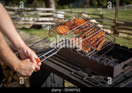 Man grilling sausages and chicken outdoors. Hands holding a barbecue grate with sausages and marinated chicken cooking over hot charcoal in a sunny Stock Photo