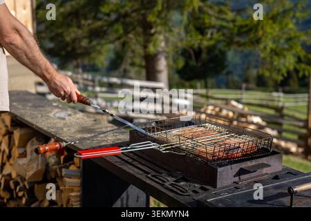 Man cooking meat on outdoor barbecue. Man uses a metal skewer while grilling meat on a charcoal barbecue in a sunny rustic mountain yard. Stock Photo