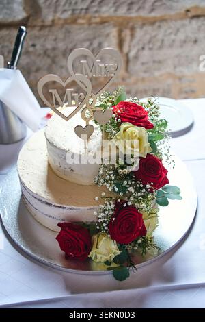 Beautiful two-tiered wedding cake decorated with fresh red and white roses, baby's breath flowers, and a wooden heart topper. Stock Photo