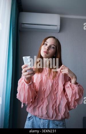 Young woman feeling warm and suffering from the heat during summer, looking at the viewer while holding a remote control to adjust her room's air Stock Photo