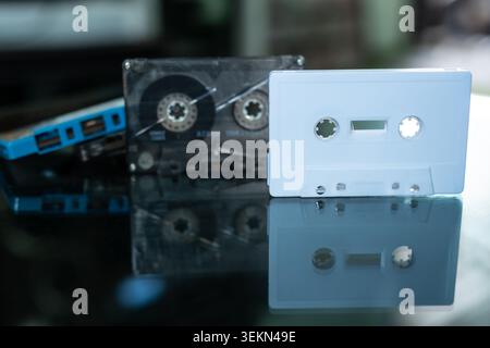 White blank cassette tape standing in front of transparent and blue vintage compact cassettes on dark reflective surface with copy space Stock Photo