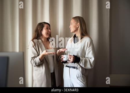 Excited businesswoman talking with female colleague standing near curtain in office Stock Photo