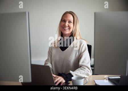 Portrait of smiling female business expert with blond hair sitting at office desk Stock Photo