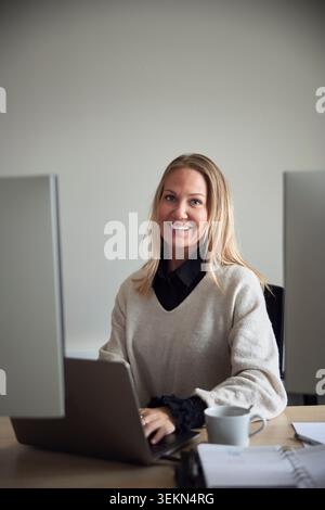 Portrait of smiling female business professional sitting with laptop at office desk Stock Photo