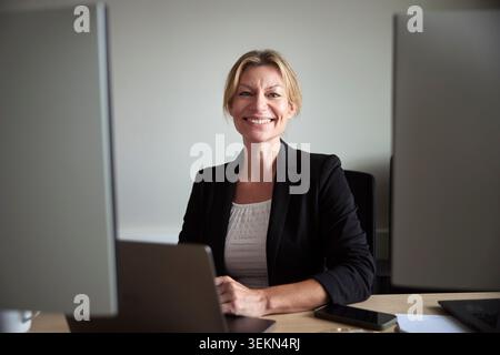 Portrait of smiling mature businesswoman sitting with laptop at desk in office Stock Photo