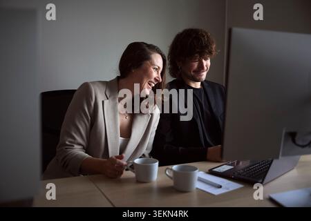 Happy businesswoman sitting near male colleague working on desktop PC in tech office Stock Photo