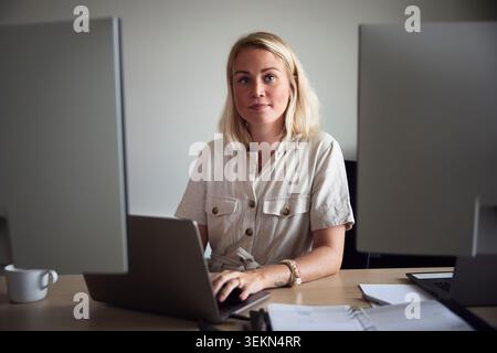 Portrait of blond young female tech professional sitting with laptop in office desk Stock Photo
