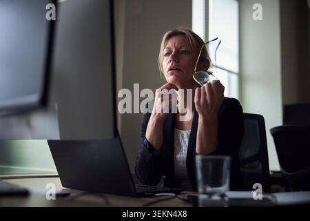Mature female tech expert holding eyeglasses looking at computer while sitting at office desk Stock Photo