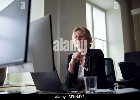 Low angle view of smiling female entrepreneur sitting with laptop and computer at office desk Stock Photo