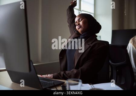 Tired female business expert stretching while sitting at office desk Stock Photo